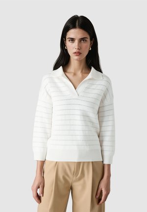 Kobieta o długich ciemnych włosach nosi biały sweter dzianinowy z dekoltem w serek, z poziomymi otworami, oraz beżowe spodnie, stoi na tle jednolitego jasnego tła.