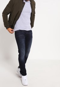 Tmavě modré džíny se štíhlým střihem, šedé tričko, olivově zelená bomber bunda, bílé tenisky. Džíny mají lehce vybledlou barvu a klasický design s pěti kapsami.