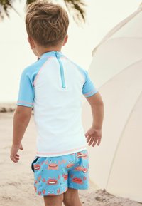 Maglietta a maniche corte in bianco e azzurro con cerniera. Pantaloni corti blu con un motivo di granchio in rosa e arancio su uno sfondo azzurro.