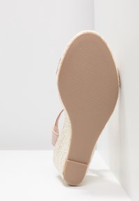 Semelle d'une sandale compensée beige à texture tissée, bride rose visible, posée contre un mur blanc en angle et un sol blanc.