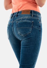 Jeans de mezclilla en azul oscuro con un diseño ajustado. Cuenta con una etiqueta de cuero marrón, bolsillos con detalles de costura y un ligero desgaste en la parte trasera.
