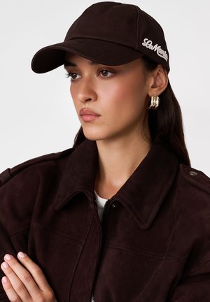 Młoda kobieta w ciemnobrązowej czapce z haftem „LeMarais”, pasującej kurtce, złotych kolczykach kołach, patrząca w lewo, z założonymi ramionami.