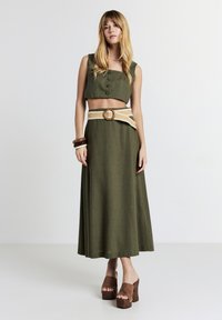 Donna con top corto buttoned verde oliva e gonna lunga abbinata, dotata di cintura intrecciata e tacchi alti marroni, in piedi contro uno sfondo bianco semplice.