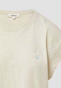 Beige katoenen t-shirt met een ronde halslijn en korte mouwen, voorzien van een pastel sterrenpatch met blauwe en gele accenten aan de linkerzijde.