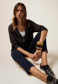 Sweat à zip noir avec des accents de perles, haut blanc, pantalons cropped bleu marine et chaussures noires à enfiler avec des clous. Plusieurs bracelets sur un poignet.
