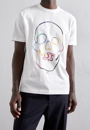 Person iført en hvid t-shirt med et farverigt, abstrakt kranieomridsdesign og mørke bukser mod en ensfarvet baggrund.