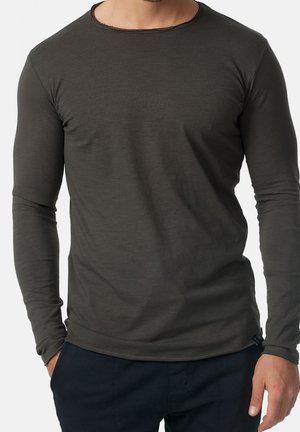 Homme portant une chemise gris foncé ajustée à manches longues et un pantalon sombre, debout avec une main dans la poche, devant un fond uni.