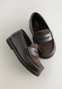 Ein Paar dunkelbraune Herrenloafers mit Wildleder-Obermaterial und Lederstreifen-Detail, schräg auf einem hellgrauen Hintergrund.
