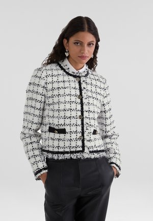 Valkoinen ja musta tweed-takki, jossa on lyhyt malli, kultaiset nappikiinnitykset, mustat taskudet ja fringed-helma.