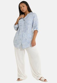 Camicia blu a fantasie paisley con maniche arrotolate, abbinata a pantaloni in lino bianchi a gamba larga e sandali chiari, su uno sfondo neutro.