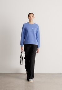 Felpa celeste con scollo tondo e logo in rilievo, abbinata a pantaloni neri a righe sottili e sneakers argentate; tiene in mano una borsa metallica.