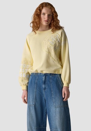 Young woman with curly red hair wears a yellow sweatshirt with lace details and loose blue denim pants against a plain background.