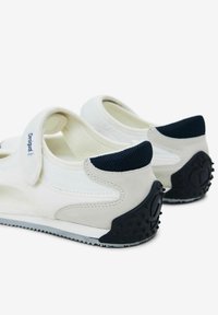 Baskets de sport blanches avec une sangle velcro, accents en maille texturée bleu marine, semelles en caoutchouc avec points de adhérence et un design épuré et fluide.