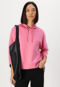 Felpa rosa con cordini, vestibilità rilassata e polsini a coste. Abbinata a una borsa nera testurizzata, su uno sfondo neutro.