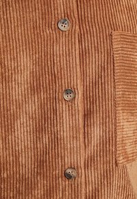 Tissu en velours côtelé brun avec des côtes verticales, trois boutons ronds beiges et une poche plaquée texturée sur le côté droit.