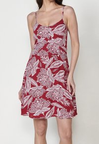 Femme portant une robe rouge sans manches avec des motifs floraux et de feuilles blancs, avec de fines bretelles et une jupe légèrement évasée.