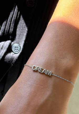Braccialetto d'argento con la parola "DREAM" in lettere maiuscole, attaccato a una delicata catena, poggiato su un polso abbronzato.