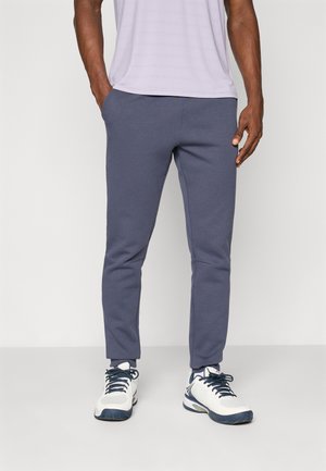 Egy férfi szürke, testhezálló melegítőnadrágot, fekete fűzőjű fehér sportcipőt és világoslila inget visel, zsebében tartja a kezét, áll.