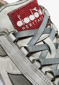 Scarpa sportiva grigia con tomaia in pelle e camoscio, caratterizzata da occhielli argentati, lacci grigi e un'etichetta in tessuto rossa con il logo "HERITAGE".