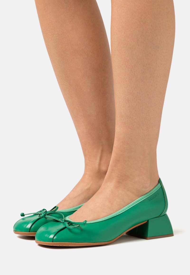 Paire de chaussures en cuir vert à talons bas, bouts arrondis et petits nœuds portées sur des jambes nues sur un fond clair.