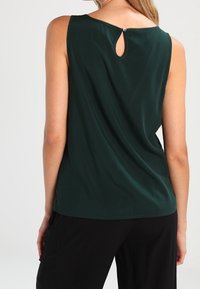 Top sans manches vert foncé avec une découpe en trou de serrure à l'arrière, texture lisse et un détail de bouton au niveau du col.