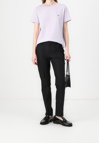 T-shirt violet clair à manches courtes avec un petit logo, associé à un pantalon noir sur mesure et des mocassins noirs, portant un sac à main noir.