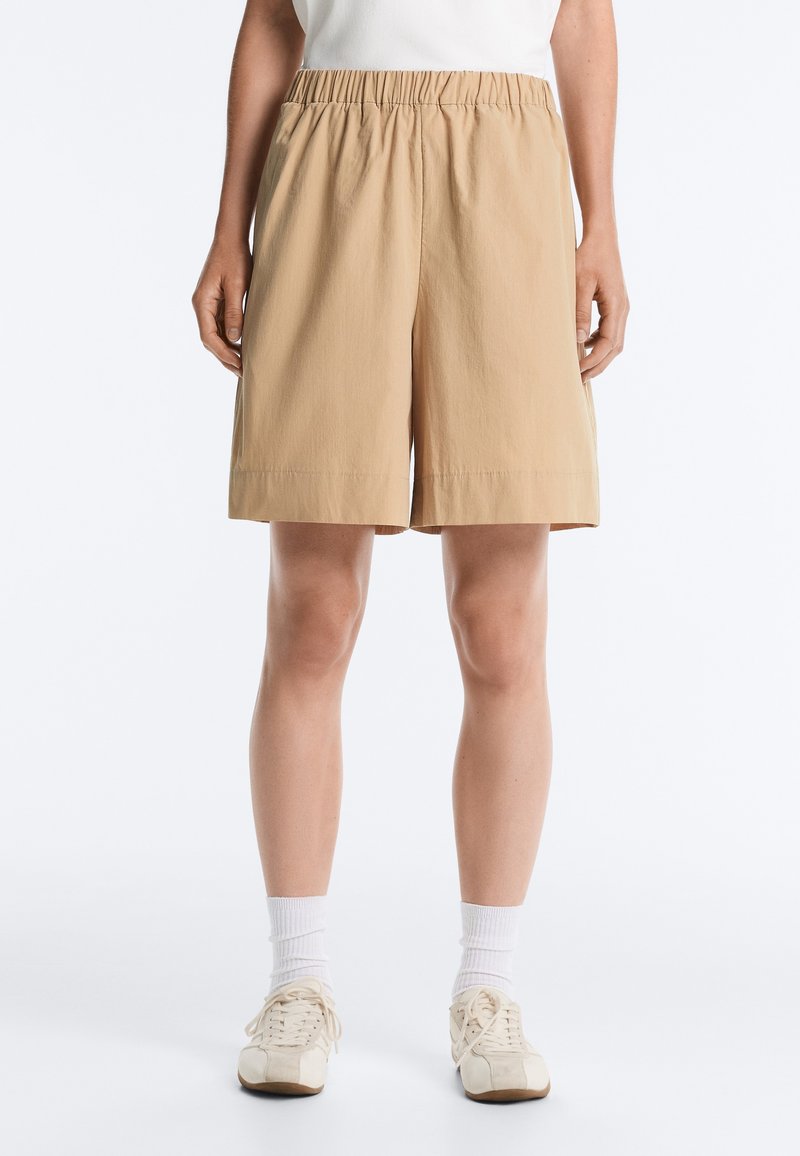 Personne portant un short beige longueur genou avec une taille élastique, des chaussettes blanches et des baskets couleur crème, debout devant un fond uni.