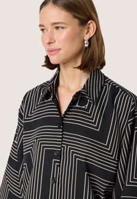 Bluză cu model negru și crem, cu un design cu butoni, având o guler mare și linii geometrice pe întreaga țesătură.