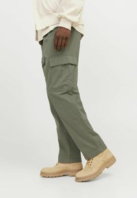 Cargo pants in olive green fabric, featuring multiple side pockets, straight fit, and paired with beige lace-up boots with textured soles.