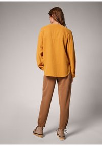 Camicia a maniche lunghe color giallo senape con orlo arrotondato, abbinata a pantaloni marroni alla caviglia e scarpe beige con finitura testurizzata.