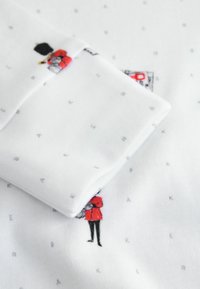 Vitt bomullstyg med små, grå bokstäver och färgglada tryckta figurer i röda rockar. Mjuk textur som visar lekfulla designelement.