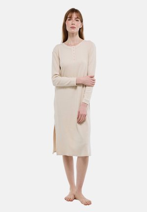 Femme debout pieds nus portant une robe midi beige à manches longues avec boutons sur le devant et fentes latérales sur un fond blanc.