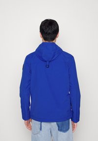 Muž oblečený v jasně modré kapuci a světlých modrých džínách s tmavou skvrnou na pravé zadní kapse, otočený zády na bílém pozadí.