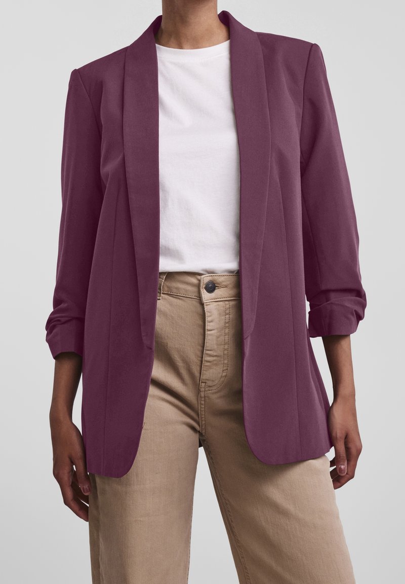 Bärnstensfärgad kavaj med sjalkrag, långa ärmar och öppen fram, kombinerad med en vit T-shirt och beige byxor. Slät textur.