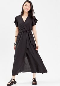 Robe noire à manches courtes volantées, ceinture à nouer et jupe fluide. Portée avec des sandales à lanières noires, créant une allure décontractée.