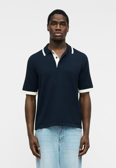 Homme portant un polo bleu marine à manches courtes avec col et bords des manches blancs, associé à un jean bleu clair, debout devant un fond gris.