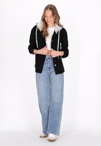 Jachetă neagră cu glugă gri, închidere cu nasturi și două buzunare frontale, combinată cu blugi albastri cu talie înaltă și adidași albi.