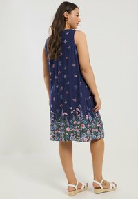 Marineblaues ärmelloses Kleid mit Schmetterlingsmuster und blumenverziertem Saum. Fließendes Design, der Stoff hat eine glatte Textur, ergänzt durch weiße Sandalen.