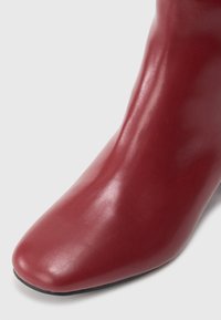 Červená kožená kotníková bota s hladkým povrchem, špičatou špičkou a elegantním designem. Podešev je černá a má minimální akcenty.