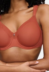 Ženska nosi modrček v rjaveči barvi z žico in majhnim srebrnim obeskom na sredini, roke pa ima prekrižane pod prsmi.