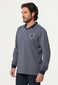 Polo-shirt met lange mouwen van donkergrijze stof met een geribde zwarte kraag met groene accenten, en een tennisclublogo op de borst.