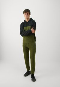 Dunkelgrüne und schwarze Kapuzenjacke mit Reißverschluss und einer Fronttasche, kombiniert mit passenden grünen Jogginghosen. Glatte Textur, sportliche Passform, Logo-Akzente.