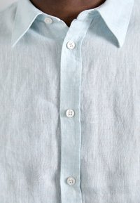 Camisa de lino azul claro con un cuello tradicional y botones blancos en la parte delantera. Tejido texturizado con un acabado natural.