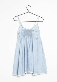 Ljusblå denimklänning med tunna band, smockad bodice, flödande kjol och fransad fåll. Hänger från en svart galge mot en vit bakgrund.