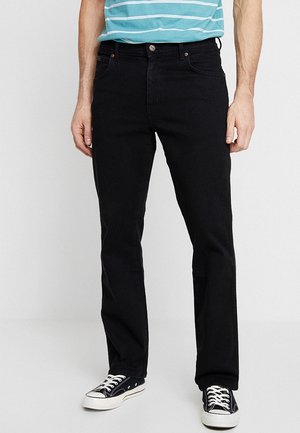 Mann trägt schwarze Jeans mit geradem Bein, schwarz-weiße Turnschuhe und ein blau gestreiftes Hemd, steht vor einem schlichten Hintergrund.