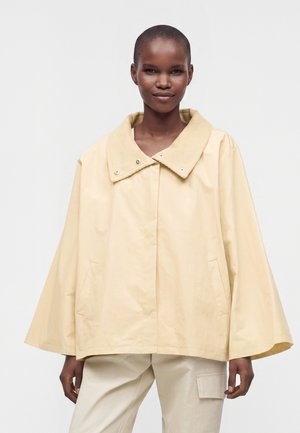 Femme aux cheveux courts portant une veste cape beige ample avec un large col et un pantalon clair sur un fond uni.