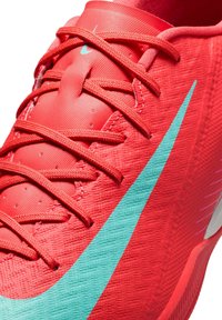 Pantof cu tălpi atletice de culoare roșie, cu un material texturat, având un logo Nike de culoare turcoaz, șireturi proeminente și o talpă amortizată.
