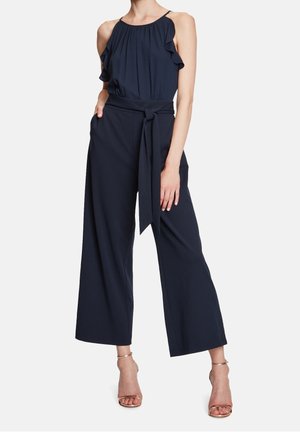 Kvinde iført en mørkeblå ærmeløs jumpsuit med flæsedetaljer, vide ben og et bindebælte i taljen, kombineret med metallic åbne hæle.