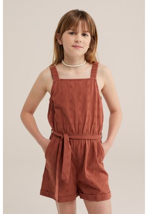 Młoda dziewczyna z jasnobrązowymi włosami, ubrana w bezrękawnikowy kombinezon koloru rdzy z paskiem do wiązania i białym koralikowym naszyjnikiem, ręce w kieszeniach.