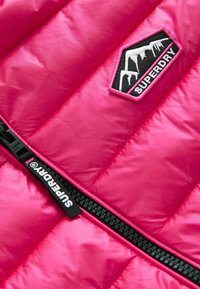 Jasne ružové vypuklé sako s prešívaným dizajnom, s čiernym zipsom a hexagonálnou čiernou a bielou nášivkou s nápisom "SUPERDRY."